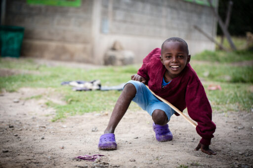 Kauernder kenianischer Junge in rotem Pullover, hellblauen kurzen Hosen und violetten Kroks