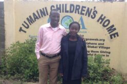 Kenianisches Paar. Er steht links (rosa Hemd, beige Hose), sie rechts (schwarzes Kleid, dunkelblaucer Mantel. Im Hintergrund ist auf einer Wand der Schriftzug "Tumaini Children's Home" zu erkennen.