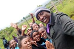 Ein Mann mit Mütze und grauem Mantel steht rechts. Links daneben sind kenianische Kinder zu sehen. Alle lächeln, auch ein paar Hände werden in die Höhe gehalten