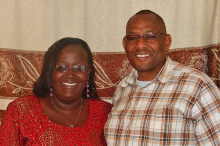 Kenianische Frau mit Brille und rotem Kleid Arm in Arm mit kenianischem Mann mit Brille und braun-weiss kariertem Hemd